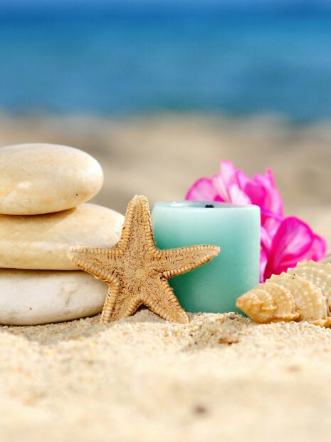 A starfish resting on sand near smooth stones and seashells with a blue candle and pink flowers