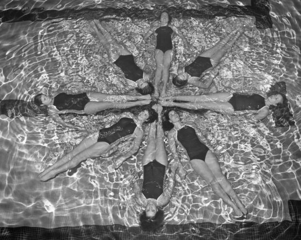 A synchronized team performing a routine floating in a pool forming a circle with their legs extended