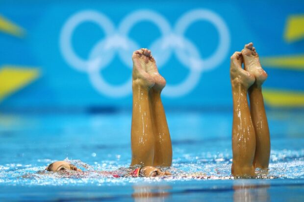 Two athletes performing synchronized swimming routine underwater with Olympic rings in background