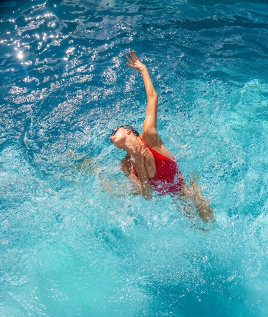 A synchronized swimmer performing an elegant move in clear blue water with sunlight reflections