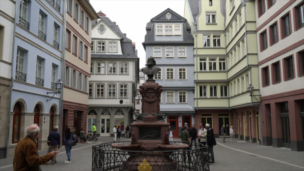Bust statue in town square surrounded by historic buildings and people walking nearby