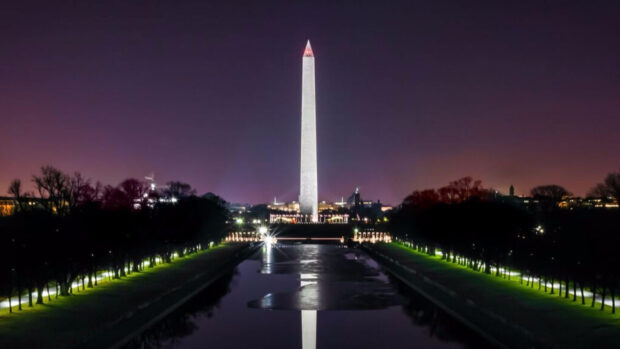 Washington Dc monument is reflected in the pool at night with illuminated trees lining the walkway