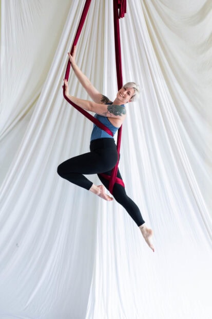 A woman performing aerial silks with tattoos and a blue top in a bright studio