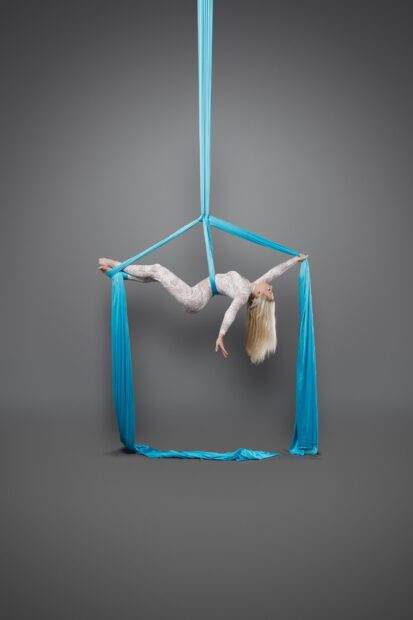 Aerial silks artist performing a suspended pose with blue silks on a gray background