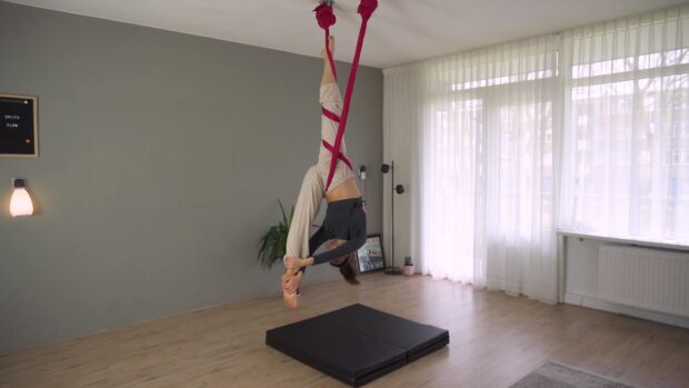 Woman practicing aerial silks exercise indoors using red fabric suspended from the ceiling