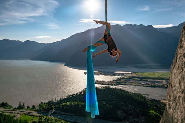 A woman performing aerial silks acrobatics hanging from blue fabric over a mountainous landscape at sunset
