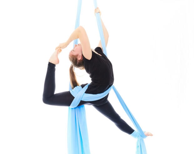 Young woman practicing aerial silks performing a flexible pose on blue fabric