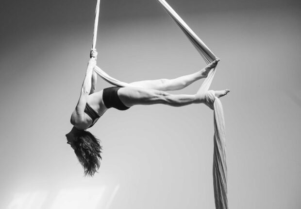 A woman practicing aerial silks hanging upside down on white fabric in a studio