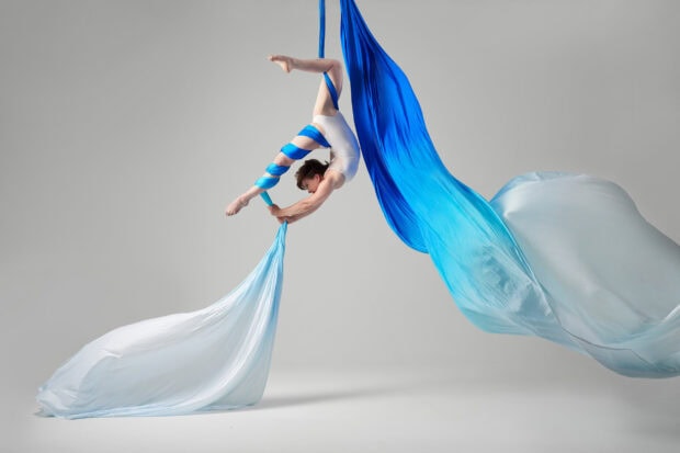 A performer demonstrating aerial silks in blue and white fabric against a gray background