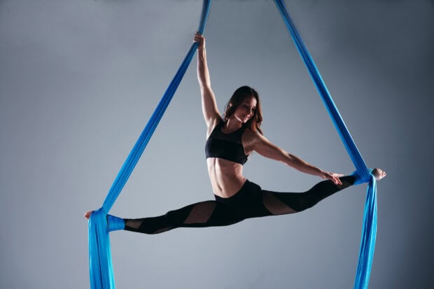 Aerial silks artist performing a split pose suspended by blue silks on a studio stage