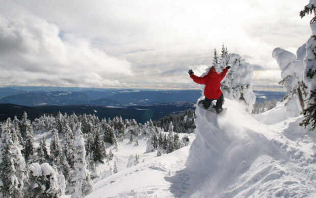 Snowboarder in red jacket riding through snowy mountain forest under cloudy sky