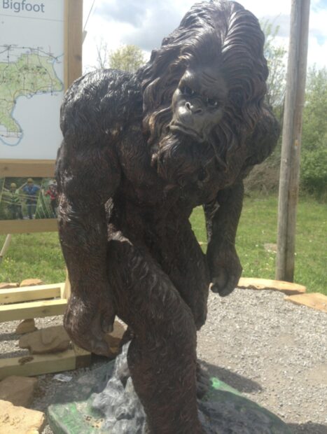 A detailed sculpture of Bigfoot standing outdoors near a wooden sign and grassy area