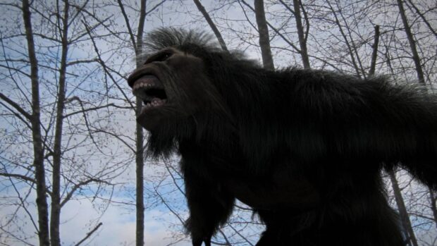A fierce bigfoot creature roaring in a forest with leafless trees and a blue sky background