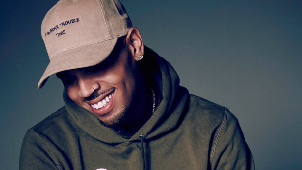 Chris Brown wearing a hat and hoodie smiling downwards in a studio setting