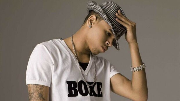Young Chris Brown wearing a checkered hat and white boxe shirt looking down and touching his hat