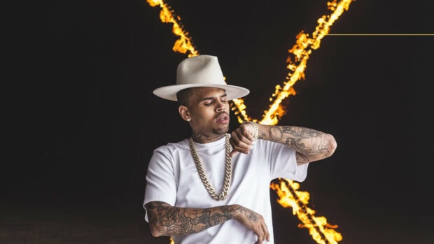 Chris Brown wearing a white hat and chain checking his watch with fire flames in the background