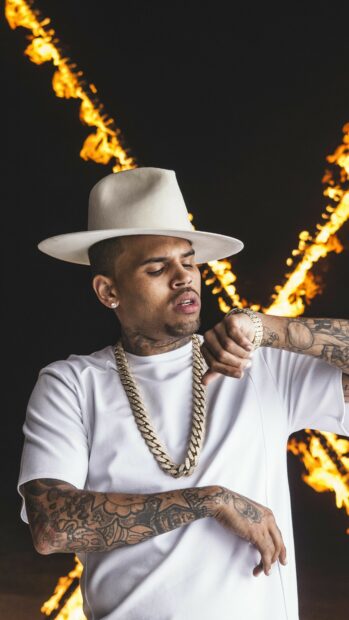 Chris Brown wearing a white hat and gold chain checking his watch with tattoos on his arms