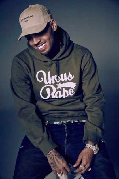 Chris Brown wearing a cap and green hoodie smiling with tattoos on his hand
