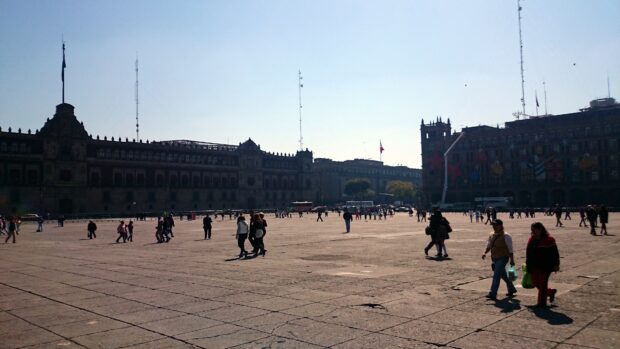 People walking across a city square with historic buildings and flagpoles in the background