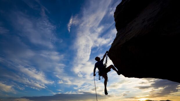 A climber ascending a rocky cliff with climbing gear against a blue sky