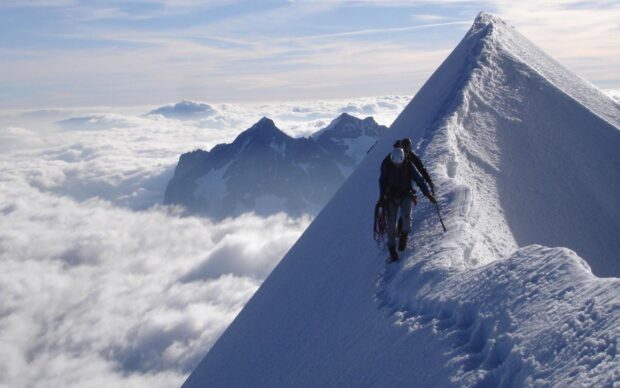 Two climbers ascending a snowy mountain ridge surrounded by clouds and distant peaks