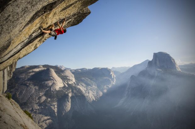 A climber wearing a red shirt ascending a steep rock face in a mountainous landscape