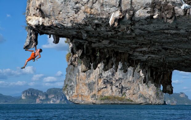 A man climbing a challenging overhanging rock formation with ocean and islands in the background