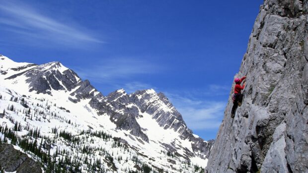 A climber in red gear ascending a steep rocky mountain face in snowy alpine terrain