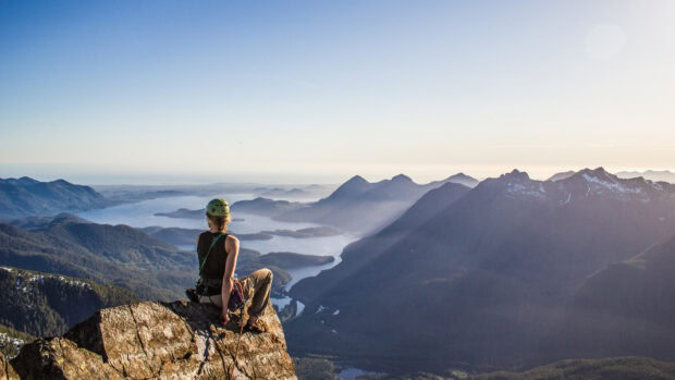 A climber resting on a rock overlooking mountains and a lake with climbing gear on