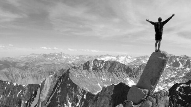 A climber celebrating on a mountain peak with arms raised overlooking a vast mountain range