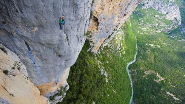 A climber scaling a steep rock face in a mountainous area with lush green forest below