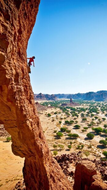 A climber scaling a steep red rock cliff in a vast desert landscape