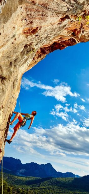 A climber ascending a steep rock formation surrounded by mountains and blue sky