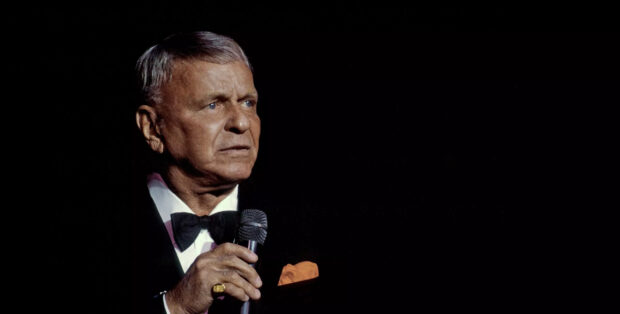 Frank Sinatra holding a microphone wearing a tuxedo on stage with a black background