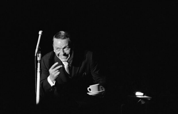Frank Sinatra sitting with a cup in hand near a microphone on stage during a performance