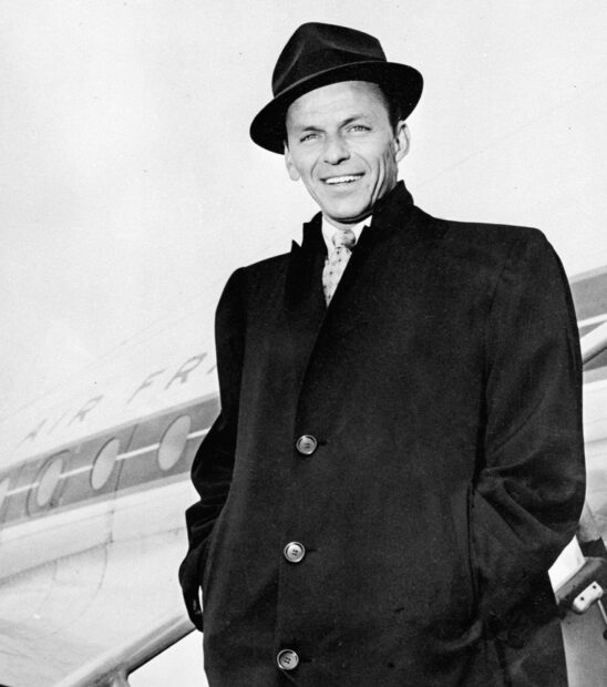 Frank Sinatra wearing a coat and hat standing near an airplane on a clear day