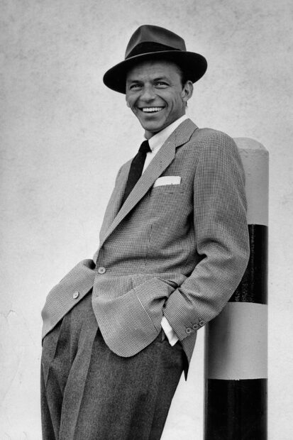 Frank Sinatra wearing a suit and hat leaning against a pole smiling confidently