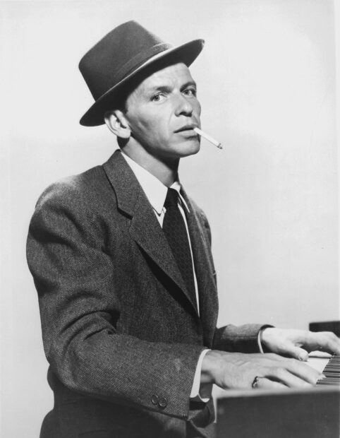 Frank Sinatra playing piano wearing a hat and suit with a cigarette in his mouth