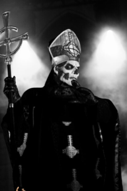 Ghost Band member dressed in ceremonial attire with skull face paint holding a staff on stage