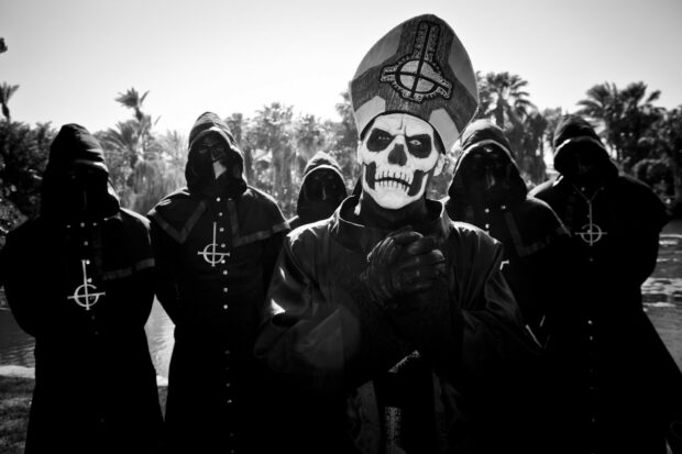 The Ghost Band members dressed in dark robes and skull face paint stand outdoors in a black and white setting