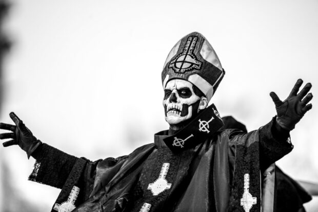 A Ghost Band member with skull face paint wearing a ceremonial robe and hat raising his hands
