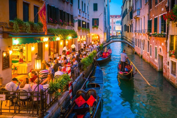 Venetian gondola navigating a narrow canal beside a lively outdoor dining area at dusk