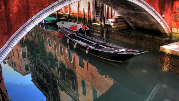 Gondola floating under a bridge with reflections of buildings in the canal water
