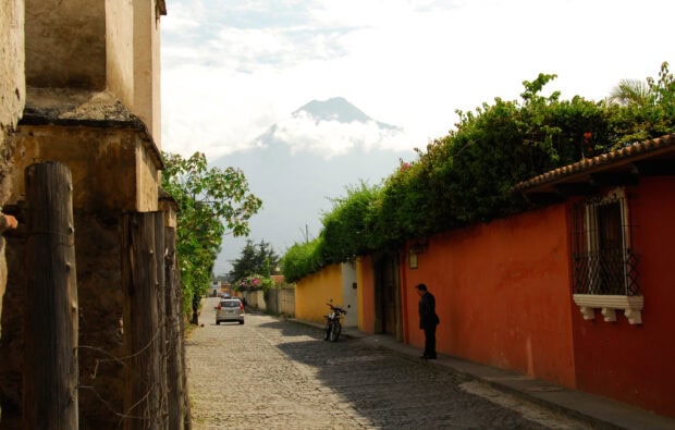 A cobblestone street in Guatemala with a man standing by an orange wall near green foliage and a distant volcano