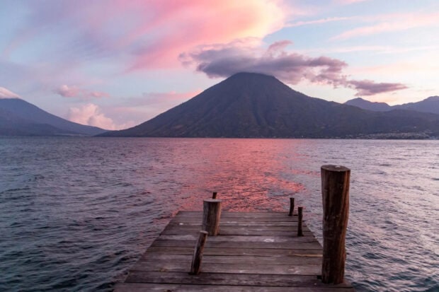 A wooden dock leading to a lake with Guatemala mountains visible in the distance at sunset