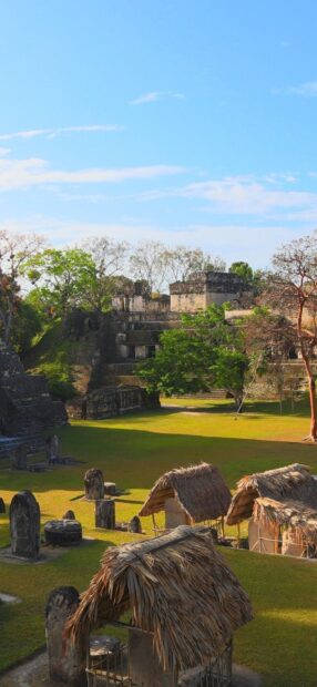 Ancient Guatemala ruins with stone monuments and thatched shelters under a clear blue sky