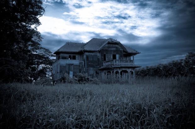 Old haunted house surrounded by tall grass under a cloudy sky at dusk