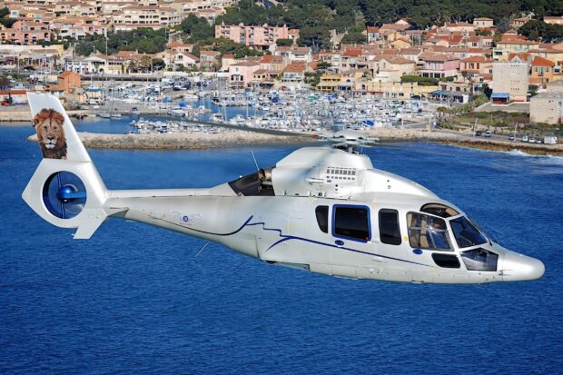 A sleek helicopter flying over the sea near a coastal town with a lion graphic on the tail