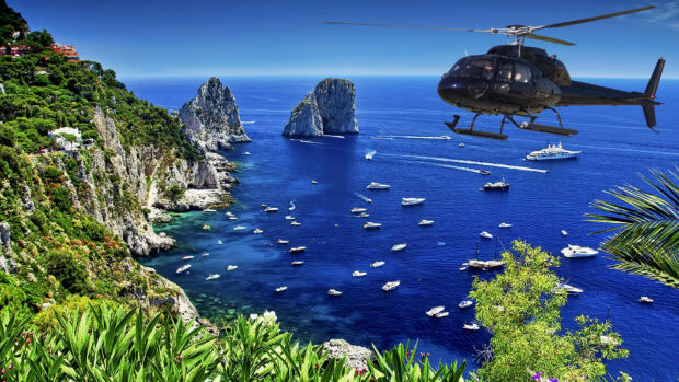 A black helicopter flying over a blue sea with rocky cliffs and many boats