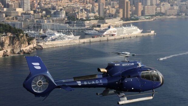 Blue helicopter flying over the bay near a coastal cityscape with yachts and buildings in the background
